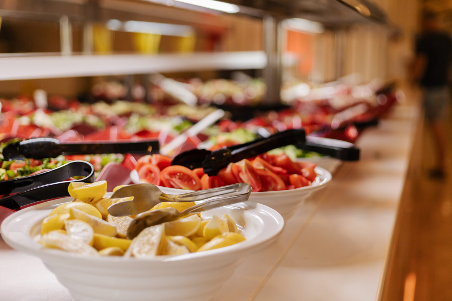 A wide selection of fresh vegetables and fruits is arranged on a salad bar. Guests can choose from bowls filled with different ingredients and use utensils to serve themselves.